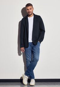Schwarze Strick-Blazer mit Taschen, weißes T-Shirt, blaue Jeans und weiße Sneakers. Das Model steht lässig an einer schlichten Wand.