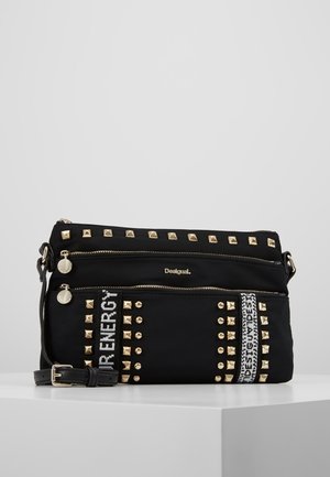 Zwarte crossbodytas met gouden studs, twee voorzakken met rits, verstelbare band en decoratieve witte tekstbanden met de tekst "YOUR ENERGY" en "DESIGUAL."
