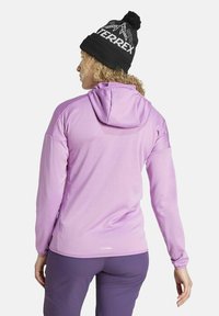 Donna con capelli ricci che indossa un berretto di maglia nero con la scritta "TERREX" e una giacca con cappuccio lavanda, vista di spalle.