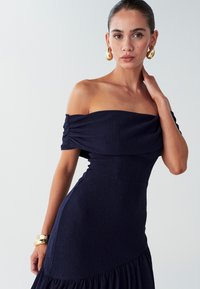 Laivastonsininen olkaimeton mekko, jossa on teksturoitu kangas, istuva bodice ja kerroksellinen röyhelöhelma. Kullanväriset käsikoru ja korvakorut korostavat ilmeen.