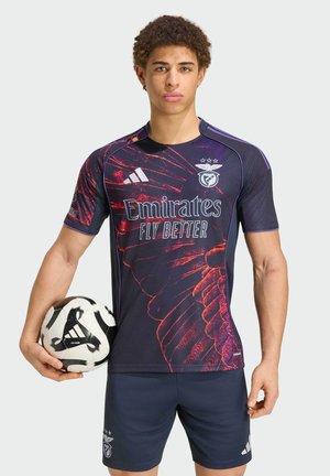 Młody mężczyzna sportowiec w ciemnoniebieskim i czerwonym stroju piłkarskim Benfiki trzyma czarno-białą piłkę nożną Adidas na białym tle.