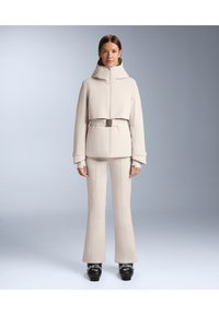 Bež zimska jakna s visokim ovratnikom, remenom i dugim rukavima, uparena s odgovarajućim širokim hlačama i crnim skijaškim čizmama. Glatka tekstura.