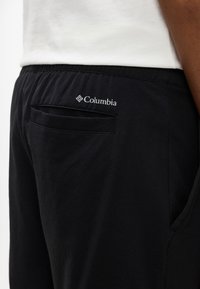 Prim-plan cu pantaloni Columbia negri cu buzunar cu fante la spate, purtați de o persoană în cămașă albă.