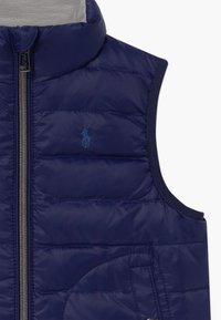 Marineblauwe gestikte mouwloze vest met een ritssluiting aan de voorkant en een klein geborduurd logo op de linkerborst.