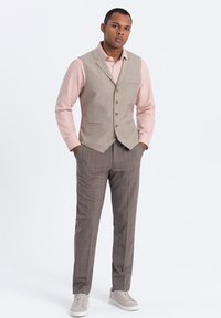 Chaleco gris claro con una tela texturizada, combinado con una camisa rosa pálido y pantalones de cuadros marrones. Zapatillas blancas completan el atuendo.