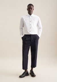 Mand iført hvid skjorte med knapper, mørke korte bukser og sorte laksko, stående med hænderne i lommerne på beige baggrund.