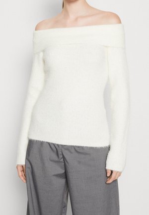 Off-the-shoulder, fitted sweater made of soft, white textured material. It has long sleeves and a wide ribbed neckline. Paired with gray trousers.