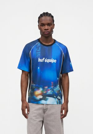 Giovane uomo con capelli intrecciati che indossa una T-shirt grafica blu con la scritta "huf équipe" e luci della città sfocate, abbinata a pantaloni grigio chiaro.
