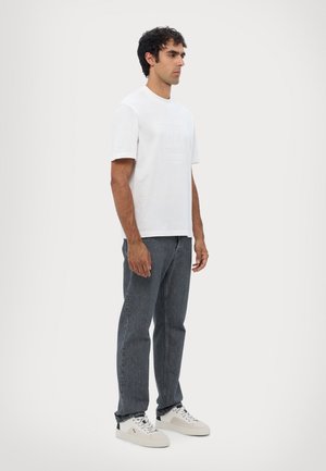 T-shirt bianca a maniche corte con logo in rilievo, abbinata a jeans in denim grigio scuro e sneakers bianche. Design semplice e casual.