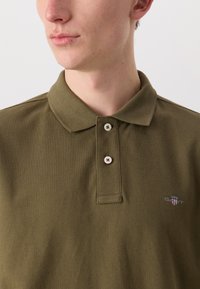 Oliven grøn polo-shirt med struktureret stof, der har en tre-knaps placket, synlige sting og et lille broderet logo på brystet.