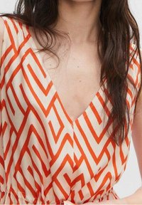 Robe blanche avec un motif géométrique orange en zigzag, featuring un décolleté en V profond et une taille froncée, fabriquée en tissu léger.