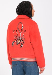 Veste en polaire rouge avec un grand motif de scorpion brodé au dos, ornée de côtes blanches et bleues sur les poignets et l'ourlet.
