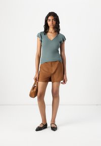 Light green short-sleeve v-neck top with ruffled sleeves, paired with brown suede shorts, black shoes with round accents, and a small brown handbag.