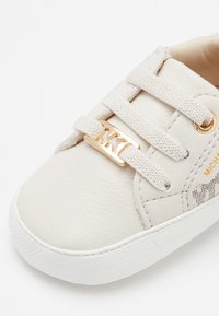 Zapatilla de cuero blanco con superficie texturizada, detalle de logo dorado, cordones elásticos y suela de goma blanca contrastante.