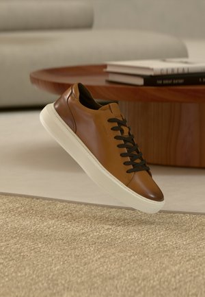 Pantof sport maro din piele cu șireturi negre și talpă albă plutind deasupra covorului bej într-un living cu masă de lemn și cărți.