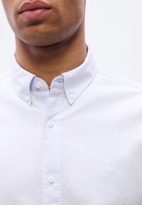 Ανοιχτό μπλε πουκάμισο με γιακά με μυτερά άκρα, διακοσμημένο με λευκά κουμπιά και μικρό ανάγλυφο λογότυπο στο στήθος. Ομαλή υφή υφάσματος.