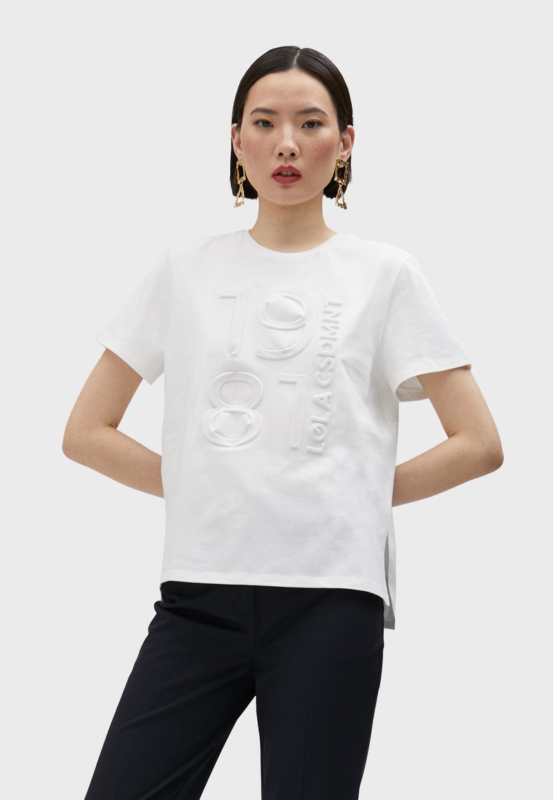 Camiseta de algodón blanca con texto gráfico en relieve "1981 LOLA CSDMNT", mangas cortas, un escote redondo y cortes laterales para un detalle adicional.