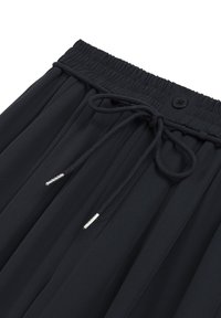 Czarne spodnie plisowane z elastycznym pasem, z silikonowym sznurkiem z metalowymi końcówkami oraz pojedynczym zapięciem na guzik.