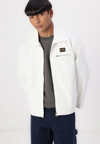 White cotton jacket with a wide collar, left chest zip pocket, and a yellow logo patch. Layered over a gray shirt, paired with blue pants.