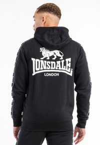 Sudadera negra de algodón, con un gran león blanco y la impresión "LONSDALE LONDON" en la parte posterior. Rayas laterales con detalles de texto repetido.