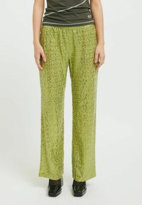 Zeleni hlače z vzorcem, ki imajo tekoč dizajn, sijočo teksturo, širok kroj nogavic in elastičen pas. Uporabljene so s sijajnim črnimi čevlji.