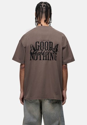 T-shirt oversize marron avec le texte graphique noir "PRÉSENTÉ PAR GOOD FOR NOTHING" et des accents de papillon au dos. Matériel en coton.