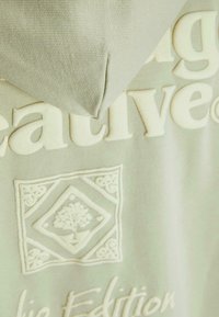 Felpa verde chiaro con testo in rilievo bianco e un emblema decorativo con un design di albero, che mette in risalto la texture e i colori in contrasto.