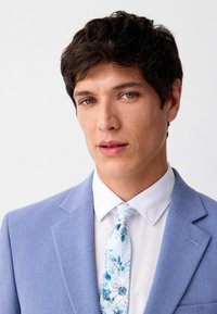 Hellblaues Sakko mit strukturiertem Finish, weißes Hemd und eine gemusterte Krawatte mit blauen Blumenmotiven. Nahaufnahme des Gesichts eines männlichen Models.