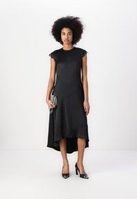 Svart satengkjole med blondekant på skuldrene, asymmetrisk hemline og høy-lav design. Modellen holder en teksturert clutch og har på seg svarte hæler.