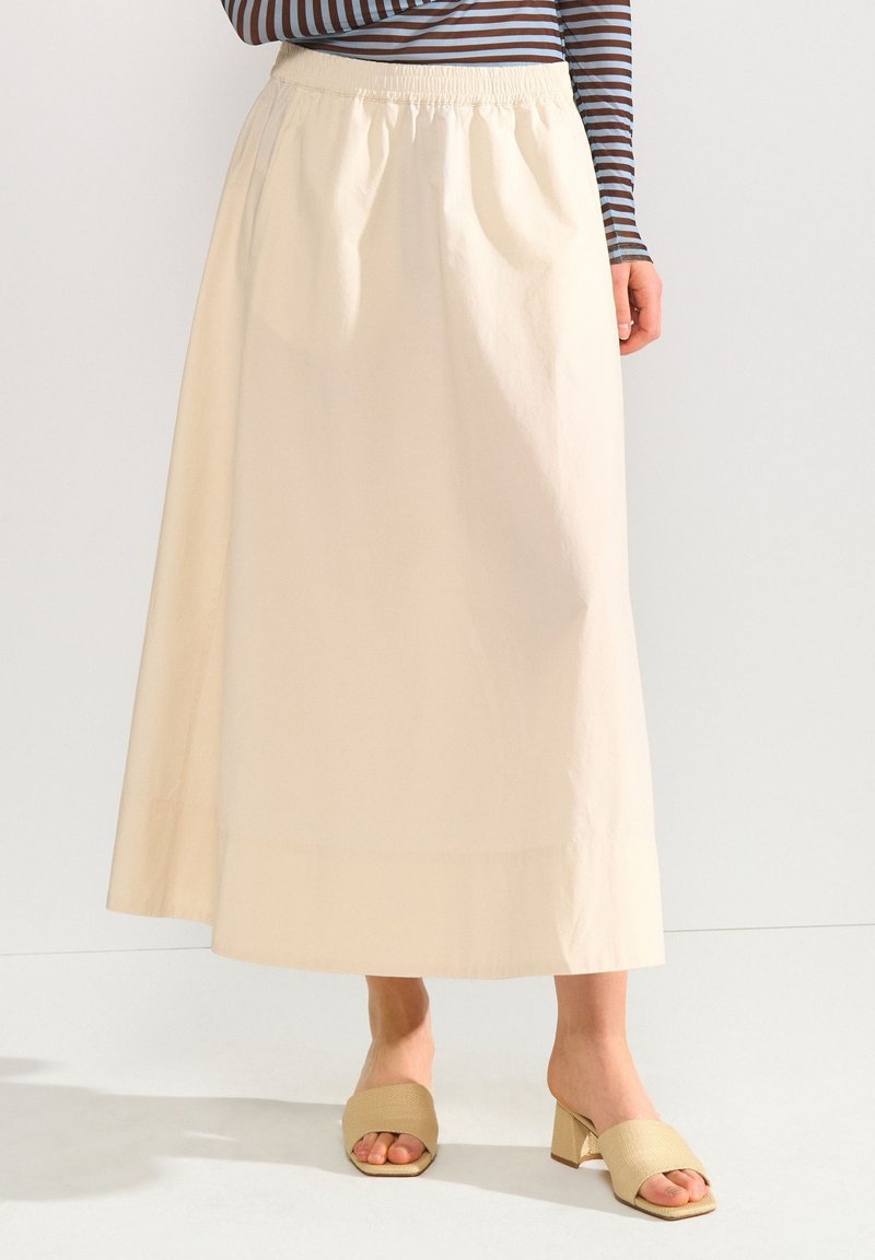 Femme portant une jupe midi beige taille élastique, associée à des sandales beiges à talons blocs ouverts et un haut rayé à manches longues.