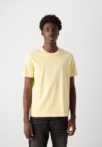 Short-sleeve yellow cotton t-shirt with round neck and small white logo on the left chest. Worn with dark jeans, standing against a plain background.