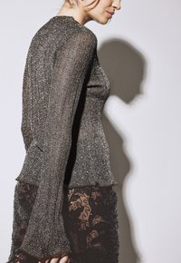 Donna che indossa un top metallico a coste con maniche lunghe e a campana e pantaloni in pizzo nero, in piedi contro uno sfondo bianco.