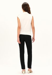 Weißes ärmelloses Oberteil mit Kragen, kombiniert mit schwarzen, eng anliegenden Hosen. Das Modell trägt hellfarbene Absätze, und die Textur wirkt glatt.