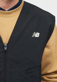 Άτομο που φοράει μία μπλούζα καφετιά με ριγέ υφή κάτω από ένα μαύρο γιλέκο με φερμουάρ και λευκό λογότυπο New Balance στο αριστερό στήθος.