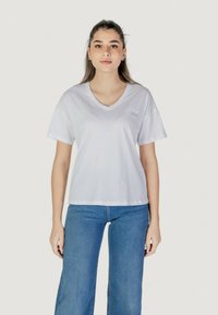 T-shirt bianco in cotone con scollatura a V e maniche corte, con un piccolo logo sul lato sinistro del petto. Abbinato a jeans a gamba larga blu.