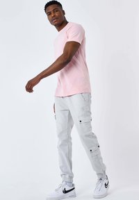 Uomo che cammina indossando una maglietta rosa, pantaloni cargo grigi chiari, sneakers bianche Nike e calzini bianchi su uno sfondo bianco semplice.
