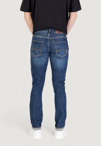 Hombre usando jeans de mezclilla azules, camiseta negra y zapatillas blancas, de pie con las manos detrás de la espalda sobre un fondo claro y liso.
