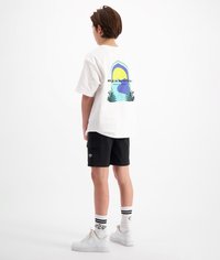 Witte t-shirt met een kleurrijk geometrisch ontwerp en tekst op de achterkant, gecombineerd met zwarte shorts en witte sneakers met zwarte accenten.