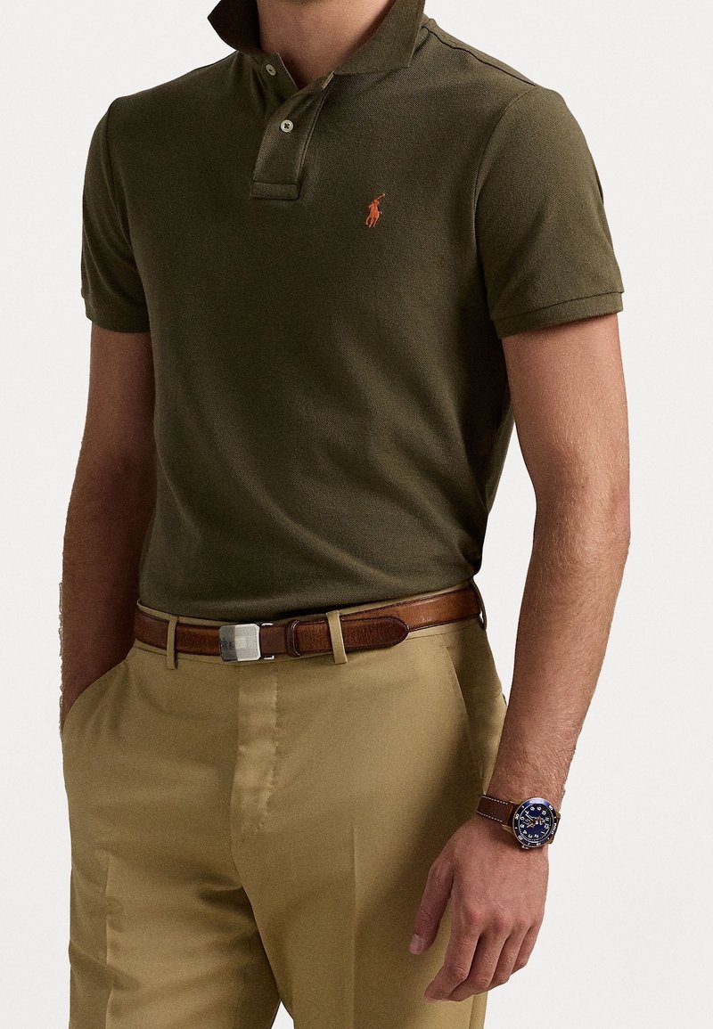 Mann trägt ein olivgrünes kurzärmliges Poloshirt, beige Hose, einen braunen Ledergürtel mit silberner Schnalle und eine braune Armbanduhr mit blauer Ziffernblatt.