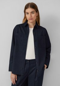 Dunkelblaues Button-up-Hemd mit vertikalen weißen Streifen, zwei Brusttaschen und langen Ärmeln. Über einem weißen T-Shirt getragen, lässige Passform.
