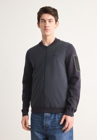 Σκούρο μπλε, ελαφρύ μπόμπερ μπουφάν με φερμουάρ μπροστά, ρίγες στο γιακά και αντίθετες σκούρες μανσέτες. Διαθέτει τσέπη με φερμουάρ στο μανίκι.