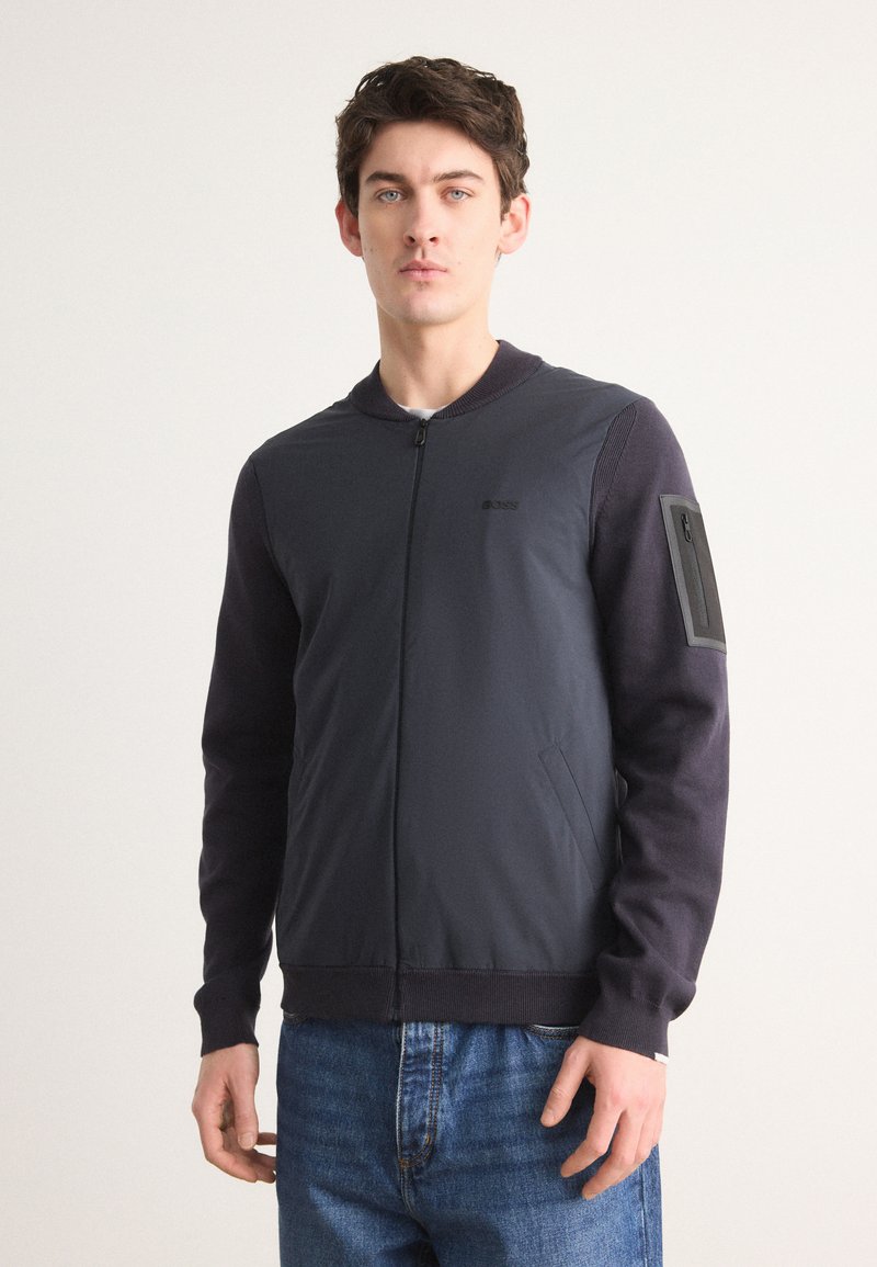 Σκούρο μπλε, ελαφρύ μπόμπερ μπουφάν με φερμουάρ μπροστά, ρίγες στο γιακά και αντίθετες σκούρες μανσέτες. Διαθέτει τσέπη με φερμουάρ στο μανίκι.