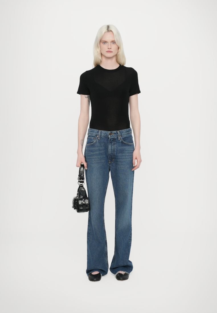 Haut noir ajusté à manches courtes, jeans bleus évasés taille haute, sac à main noir avec quincaillerie argentée, chaussures plates noires, fond blanc uni.