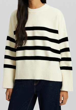 Pull crème avec de larges rayures noires horizontales. Présente un col rond côtelé et des poignets côtelés. Texture douce, coupe décontractée et épaules tombantes.