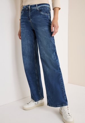 Des jeans en denim bleu à jambe large présentent un délavage usé avec de subtils frottis, un design classique à cinq poches et des coutures contrastées. Associés à des baskets blanches.