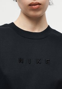 Κοντινό πλάνο ενός ατόμου που φοράει μαύρο πουκάμισο με στρογγυλή λαιμόκοψη, με ψηλά κεντημένα μαύρα γράμματα "NIKE" στο στήθος.