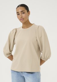 Beige puff-sleeve top made from textured fabric, featuring a round neckline and gathered cuffs, paired with light blue jeans.