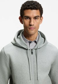 Giovane uomo con capelli ricci scuri che indossa una felpa con cappuccio grigio chiaro e cerniera sopra una camicia con colletto, rivolto in avanti con espressione neutra.