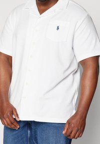 Hombre vestido con una camisa de botones de manga corta blanca con un logo de jugador de polo azul en el bolsillo del pecho y jeans azules.