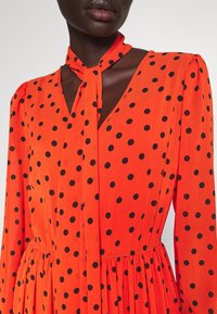 Personne à la peau foncée portant une robe orange vif à pois noirs, manches longues, col en V et une cravate assortie au cou.
